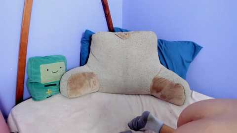 A video showing a beige plush chair with a grey paw print on the seat, a green plush figure with a star, and a blue pillow on a white bed in a corner, with a hand holding a grey object.