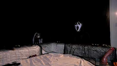 Video of a dimly lit room at night, featuring a large, eerie mask with a wide, open mouth and prominent eyes hanging from a wooden beam. The background is dark, emphasizing the creepy atmosphere.