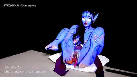 Video of a blue-skinned, naked woman with pointy ears, sitting on a white cloth, legs spread, against a black background, revealing her genitals.