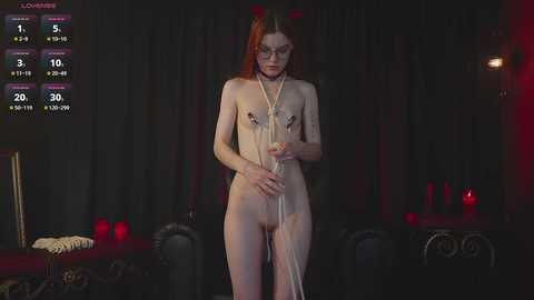 A video of a nude, slender woman with fair skin and long red hair, standing in a dimly lit, red-lit room, wearing only a string of pearls, surrounded by black curtains and red candles.