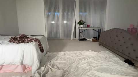 Video of a minimalist bedroom with a white bed, leopard-print blanket, beige carpet, and sheer white curtains. The room features a grey upholstered headboard, a black side table, and a pink lamp.