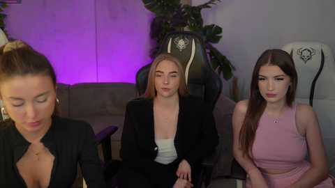 Media: Video of three young women in a dimly lit room: one in a black top, another in a black blazer, and a third in a pink crop top. Gaming chairs, green plants, and purple lighting background.