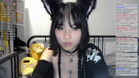 Media: A young Asian woman with black hair, wearing cat ears and a black choker, sits on a bed, surrounded by plush toys. She's in a dimly lit room with a chat window visible on the right.