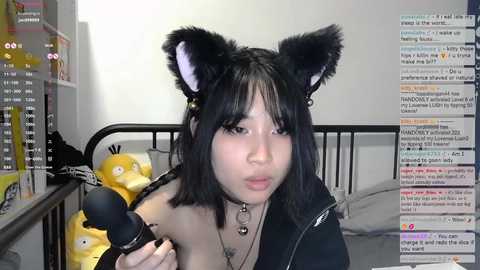 A video of a young Asian woman with black cat ears, wearing a black hoodie, making a kissy face into a microphone in a messy room with a bed, stuffed toys, and a computer screen displaying chat logs.