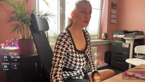 Video of a blonde woman in a black-and-white patterned top, sitting in a black chair, in an office with pink walls, a window, a desk, and a potted plant.