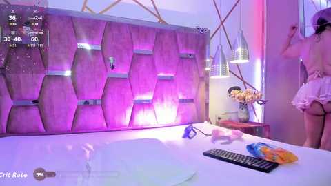 Video of a modern bedroom with a pink, geometric headboard, pink and purple ambient lighting, and a woman in a white dress standing beside a bed with a remote control and a box of tissues.
