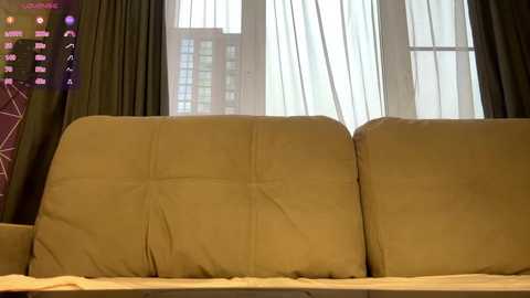 Video of a beige sofa with tufted cushions in a modern living room, featuring sheer white curtains and dark green drapes in the background, with a cityscape visible through the windows.