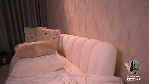Video of a cozy bedroom with a plush white bed, beige pillows, and textured cream walls. A social media icon with a heart emoji and \"Mariana\" is displayed in the bottom right corner.