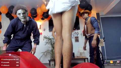 Media: Video of a woman in a white dress standing with her back to the camera, surrounded by two men in Halloween costumes. Orange balloons and decorations adorn the background.