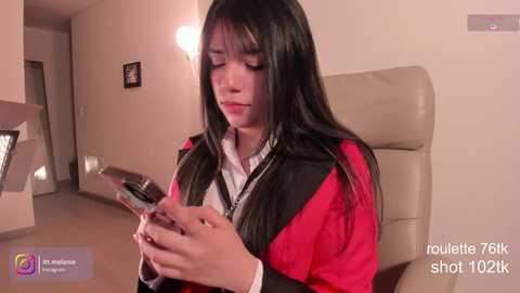Media: Video of a young Asian woman with long black hair, wearing a red and black jacket, intensely focused on her smartphone in a modern, beige-toned hallway.