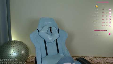 Video of a light blue gaming chair with a cushioned backrest and armrests, placed in a softly lit room with a beige carpet and a silver exercise ball. A gaming app screen displays stats and options in the background.