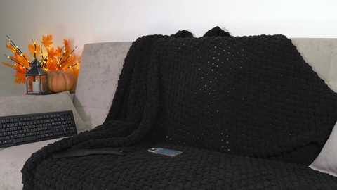 Video of a cozy living room scene with a black knitted blanket draped over a beige couch. An orange pumpkin and candle centerpiece is visible in the background, creating a warm, autumnal atmosphere.