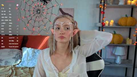 Video of a blonde woman with elf ears and white face paint, wearing a sheer white top, in a cozy room with Halloween decorations and lit candles.
