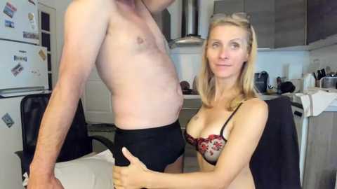 Video of a slim, shirtless man wearing black briefs and a blonde woman in a floral bra, standing in a modern kitchen with white cabinets and black countertops.