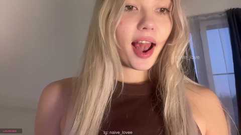 Video of a young, fair-skinned woman with long, blonde hair, wearing a brown sleeveless top, sticking out her tongue. Background shows a window with a blue curtain.
