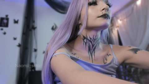 Video of a pale-skinned woman with long, pastel purple hair, wearing a grey tank top. Her face and neck are adorned with intricate black tattoos resembling tree branches. Background features black butterflies and fairy lights.