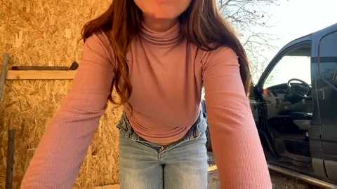 Video of a woman with long brown hair, wearing a pink long-sleeve top and light blue jeans, leaning against a black car parked next to a wooden shed.