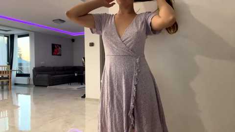 Media: Video of a woman with fair skin, long brown hair, wearing a light purple, V-neck, ruffled dress, standing in a modern living room with beige walls, a gray sectional sofa, and purple LED strip lighting.