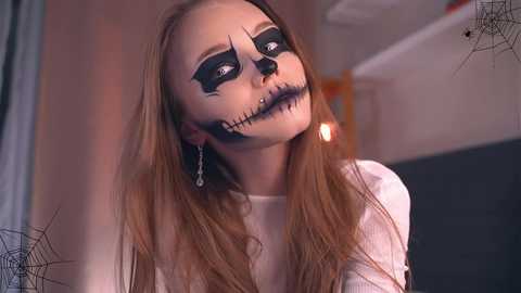 Media: Video of a young woman with pale skin, long straight hair, and a white dress, wearing a spooky Halloween mask with black makeup and a stitched mouth. Background includes a spider web and dim lighting.