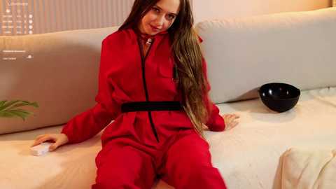 Video of a woman in a bright red jumpsuit, sitting on a cream couch, with a black bowl beside her and a green plant in the background.