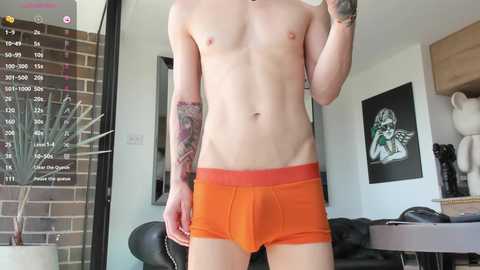 Video of a slender, light-skinned man with tattoos, wearing tight, bright orange briefs, standing in a modern living room with a brick wall, black leather couch, and framed art.