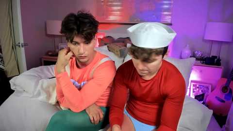 Video of two men in costumes: one in a red shirt, white hat, and blue shorts, the other in a pink shirt, green pants, and white hat. They sit on a bed in a dimly lit room with purple and pink lighting.
