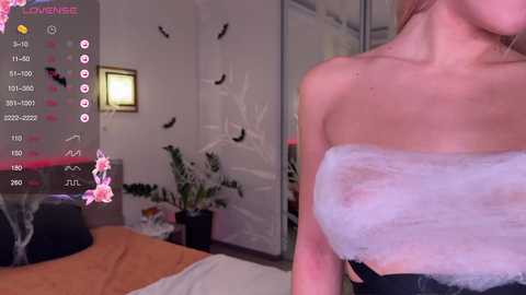 A video of a woman with fair skin, wearing a fluffy white towel over her chest, in a dimly-lit bedroom with a mirror, plants, and a bed.