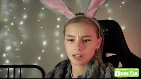 Video of a young woman with fair skin and blonde hair, wearing a pink bunny ears headband, fur coat, and distressed look. Background features sparkly lights and a black chair.