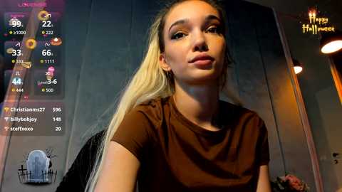 Video of a young, fair-skinned woman with long blonde hair, wearing a brown t-shirt, standing indoors, with a digital screen displaying weather and social media information in the background.