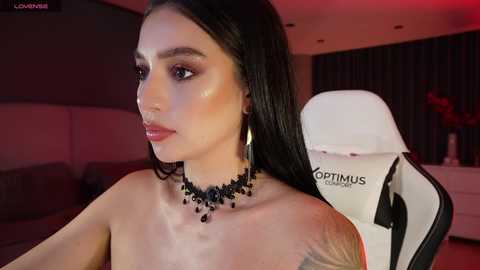 Video of a light-skinned woman with long black hair, wearing a black choker with dangling beads, sitting in a white gaming chair, indoors with dim lighting.