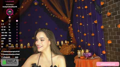 Video of a smiling young woman with light skin and brown hair in a black dress, surrounded by Halloween decorations including orange pumpkins and candles.