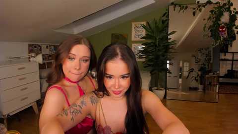 Video of two women, one with tattoos on her chest, standing in a cozy, well-decorated room with green walls, wooden floor, and plants.
