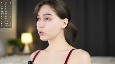 Video of a young, light-skinned woman with straight, dark hair tied back, wearing a red strapless top. Background features a blurred, dark curtain and a lamp.