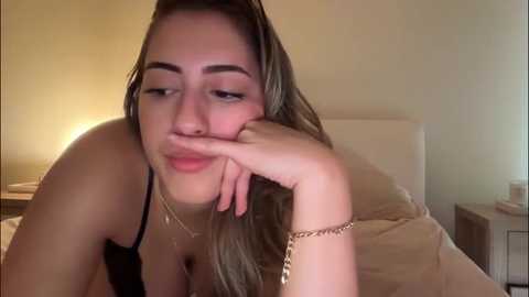 Video of a young woman with fair skin, long brown hair, and light makeup, resting her chin on her hand, wearing a black bra, in a dimly lit bedroom.