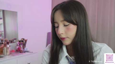 Video of a young Latina woman with long, dark hair, wearing a white shirt, applying makeup in a brightly lit room with a vanity table and mirror.