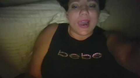 Media: Video of a woman with light skin, dark hair, and full lips, wearing a black sleeveless shirt with \"Babe\" in white letters. She is lying on a bed with beige pillows and sheets, giving a sultry look.