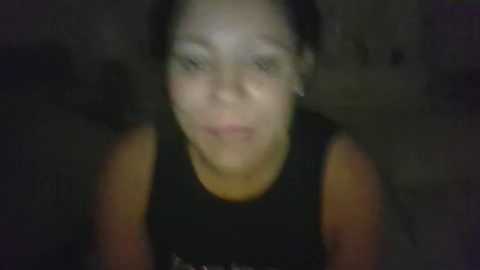 Media: A blurry video of a young woman with medium skin tone, dark hair, and a black tank top, smiling slightly. The background is indistinct and dark.