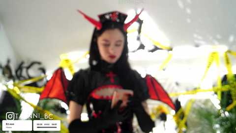 A video of a young woman in a black, red, and yellow devil-themed cosplay costume with red horns, wings, and a high-collared outfit, standing in a brightly lit room with yellow streamers.