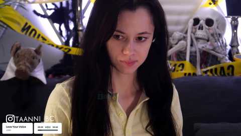 Video of a young woman with long black hair, wearing a yellow button-up shirt, standing in a spooky room with Halloween decorations, including a skeleton, a stuffed dog, and yellow caution tape.