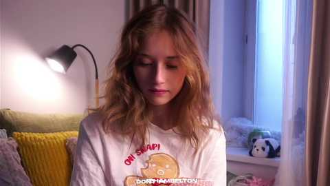Video of a young woman with long, wavy blonde hair, wearing a white t-shirt with a cartoon dog graphic, standing in a softly lit bedroom with a yellow couch, a lamp, and a window.
