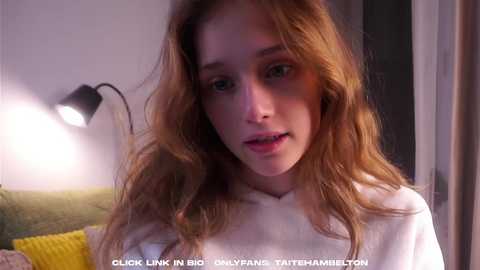 Media: A video of a young Caucasian woman with wavy, light brown hair, wearing a white sweater, standing in a dimly lit room. The background shows a green couch and a black lamp.