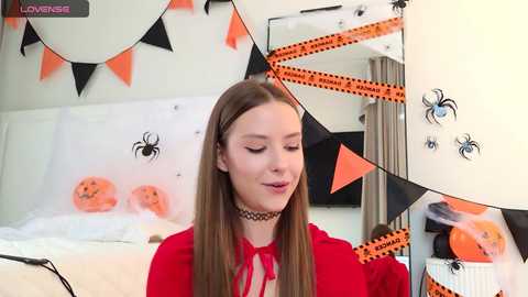Video of a young woman with straight, long brown hair, wearing a red top and black choker, surrounded by Halloween decorations including orange triangular flags, black spider cutouts, and pumpkin banners.