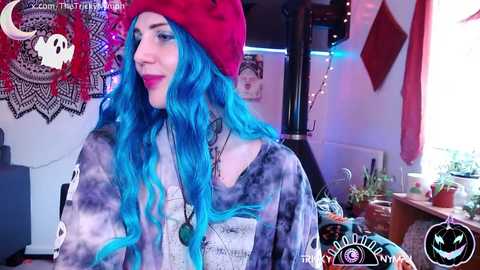 Video of a young woman with vibrant blue hair, wearing a red beanie and a tie-dye top, standing in a cozy, dimly lit room with mystical decorations.