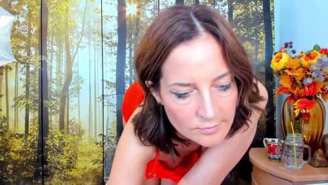 Video of a fair-skinned woman with shoulder-length brown hair, wearing a red top, looking contemplative with hands on her head. Background features a forest mural and a flower vase.