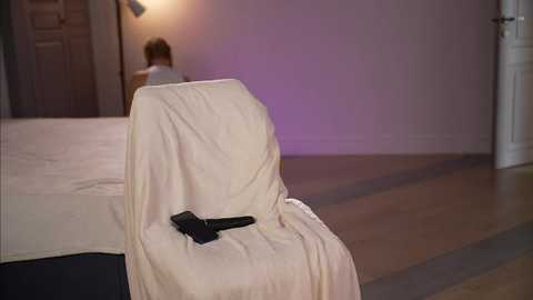 A video shows a beige chair covered with a white sheet, with a black handgun on the seat, in a dimly lit room with purple walls and wooden floor.