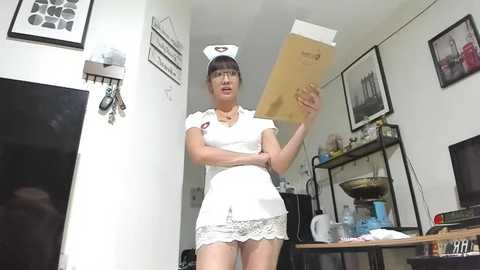 Media: Video of a petite Asian woman in a nurse outfit, holding a wooden sign, in a cluttered, minimalist room with framed artwork and kitchen items.