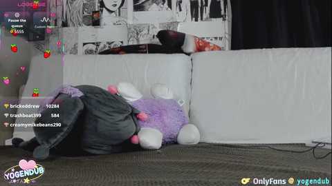 Video of a purple teddy bear with pink eyes and ears, lying on a white sofa. Background includes a black curtain and a framed manga page. Overlay text includes usernames and emojis.