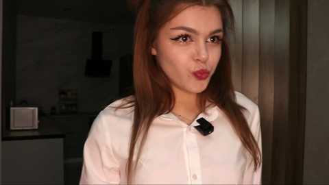 Video of a young woman with long, straight brown hair, wearing a white blouse, puckering her lips, indoors with modern kitchen background.