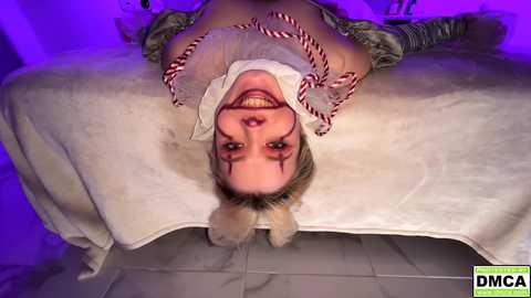 Video of a woman with blonde hair, wearing a white shirt and red bow tie, lying on a white blanket, upside down, smiling.