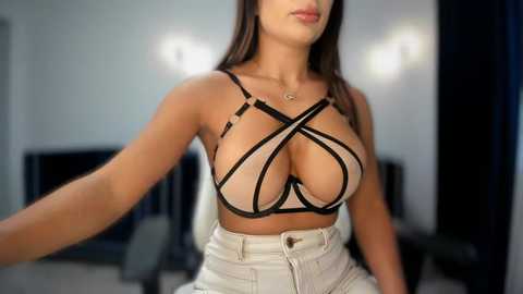 Video of a curvy, light-skinned woman with long brown hair, wearing a revealing black and white strappy bralette that accentuates her large breasts, paired with high-waisted white pants, in a modern, dimly lit room with white walls and a TV.